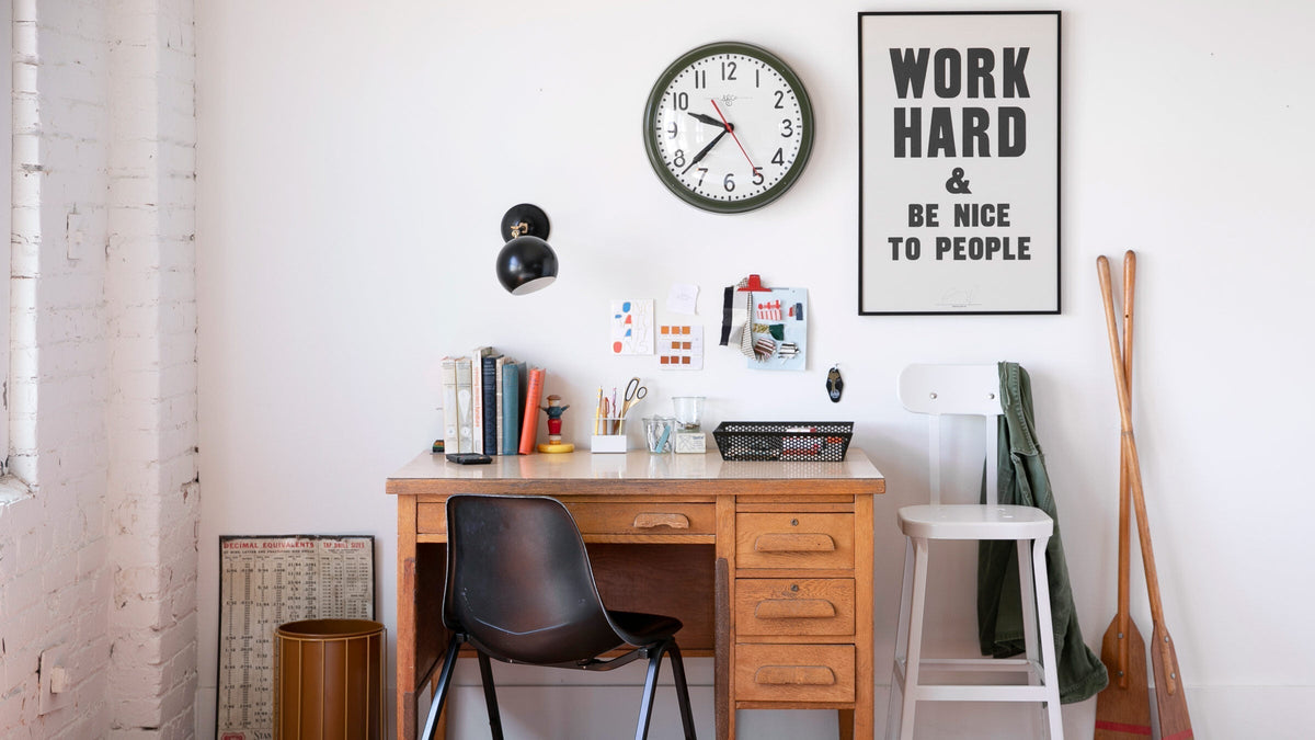 Organized home office desk with wooden drawers, black chair, wall clock, and poster reading ‘WORK HARD AND BE NICE TO PEOPLE’