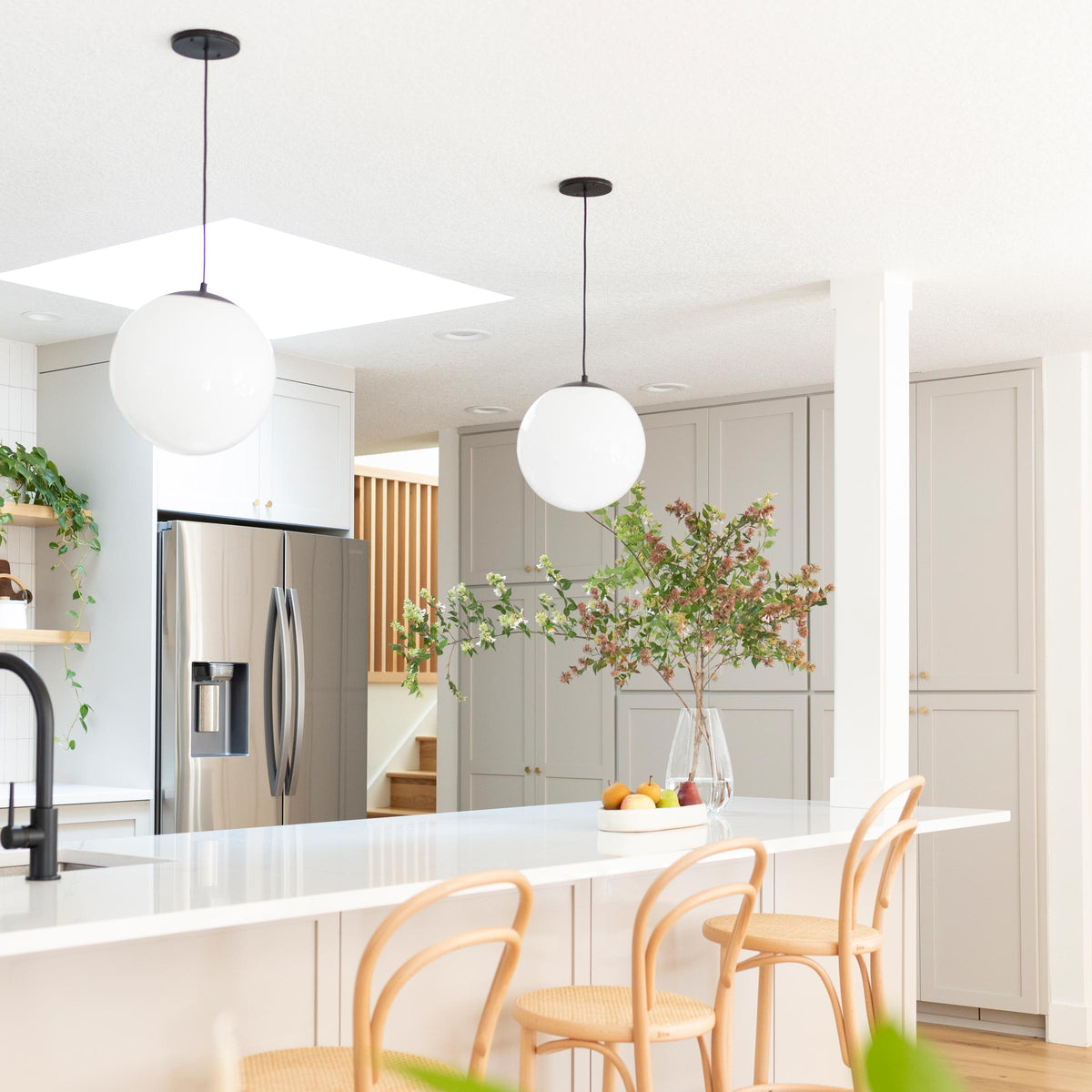 Kitchen island with two white globe pendant lights, marble countertop, woven wood bar stools, and a glass vase of branches in a bright modern kitchen