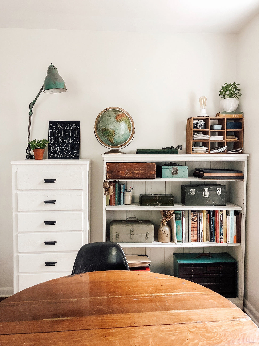 Shelf and dresser styled with a vintage green lamp and globe, alphabet chalkboard, potted plants, books, and storage boxes against a white wall