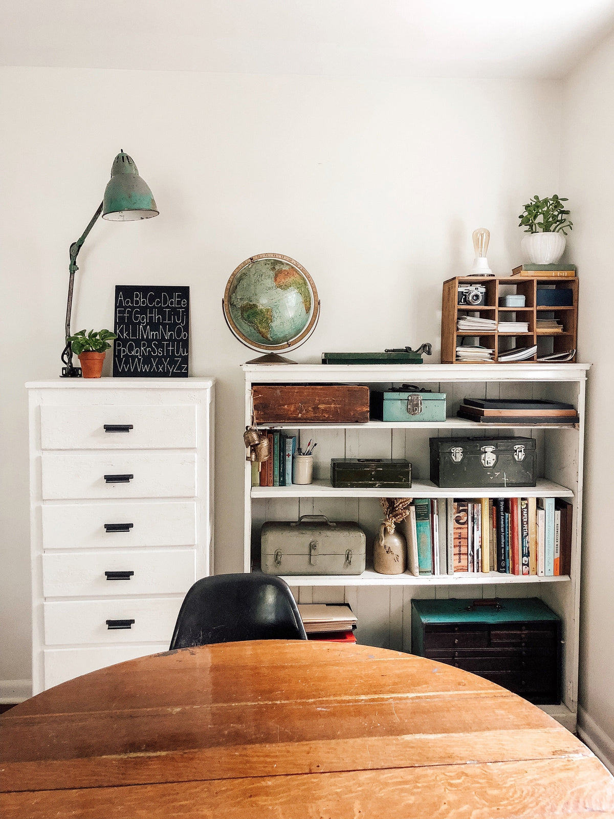 Shelf and dresser styled with a vintage green lamp and globe, alphabet chalkboard, potted plants, books, and storage boxes against a white wall