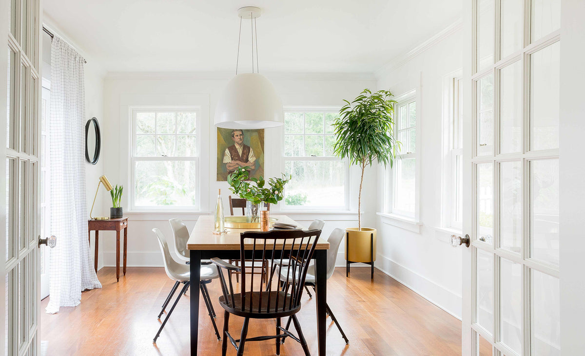Sunlit dining room with wooden table and mixed chairs, large white pendant light, and a tall potted tree by the windows