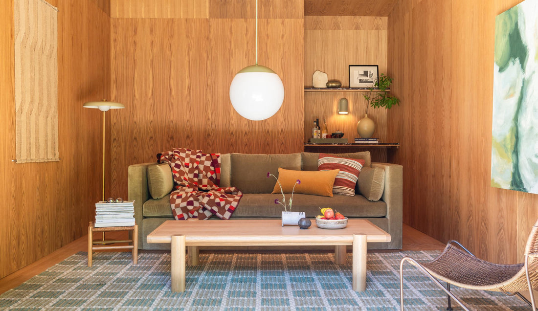 Living room with olive-green sofa, patterned throw, wood-paneled walls, round white pendant light, and low wooden coffee table
