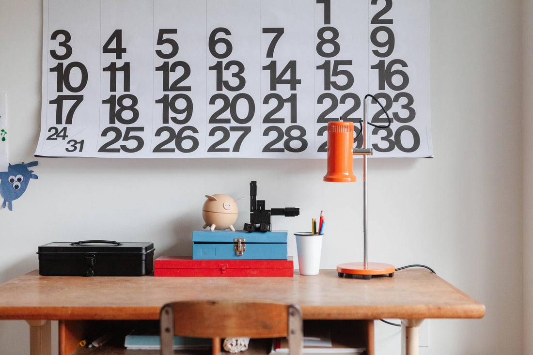 Orange Joe Task lamp standing on a wooden desk beside stacked boxes, a pencil cup, and a large black-and-white calendar on the wall
