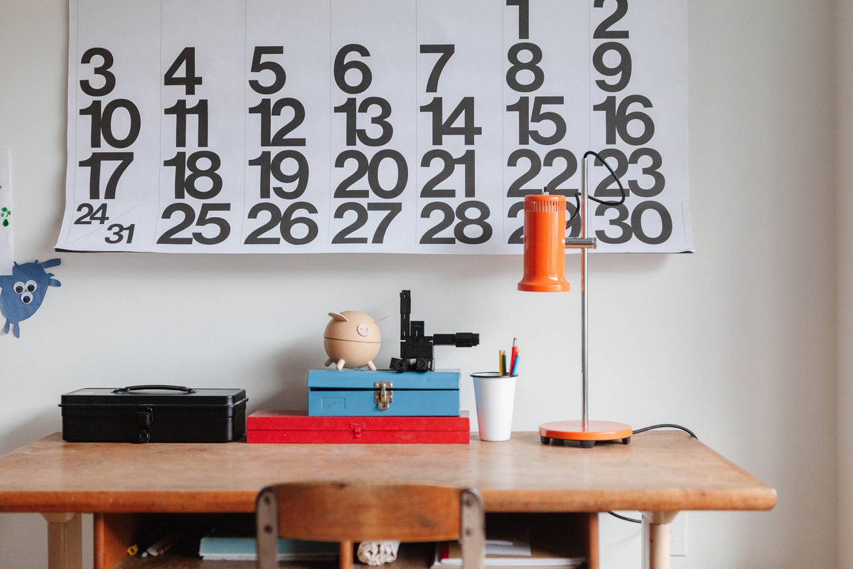 Orange Joe Task lamp standing on a wooden desk beside stacked boxes, a pencil cup, and a large black-and-white calendar on the wall