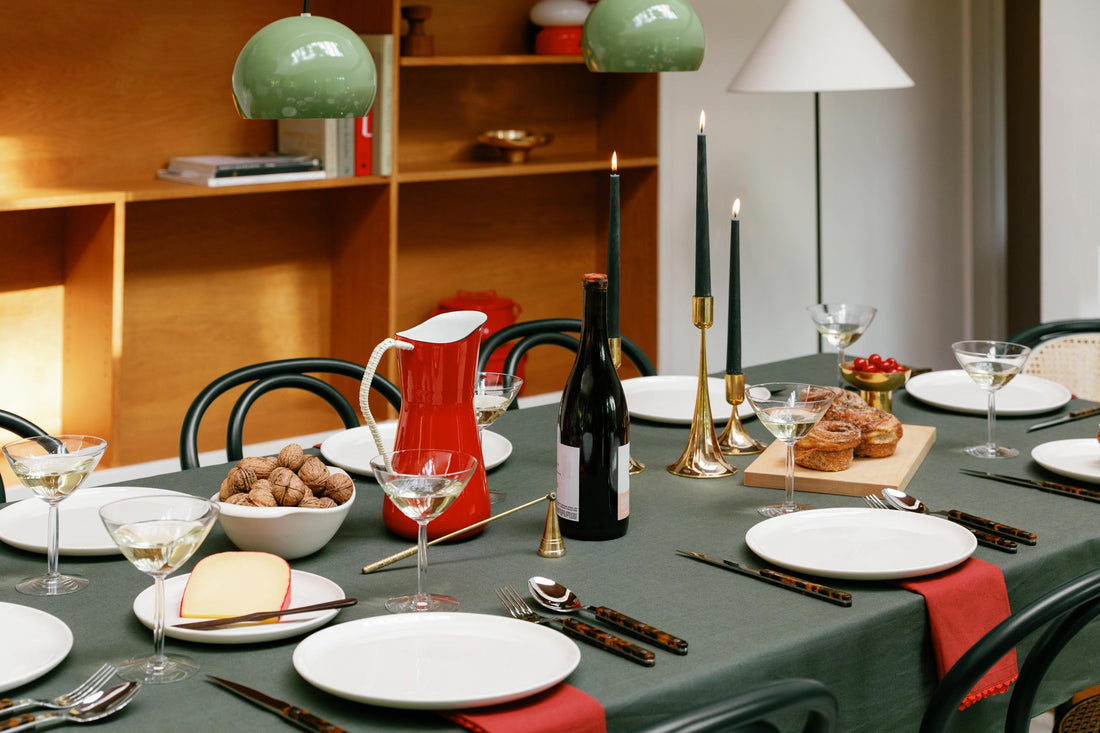 Holiday dining table set with green tablecloth, white plates, red napkins, red pitcher, wine bottle, lit black taper candles