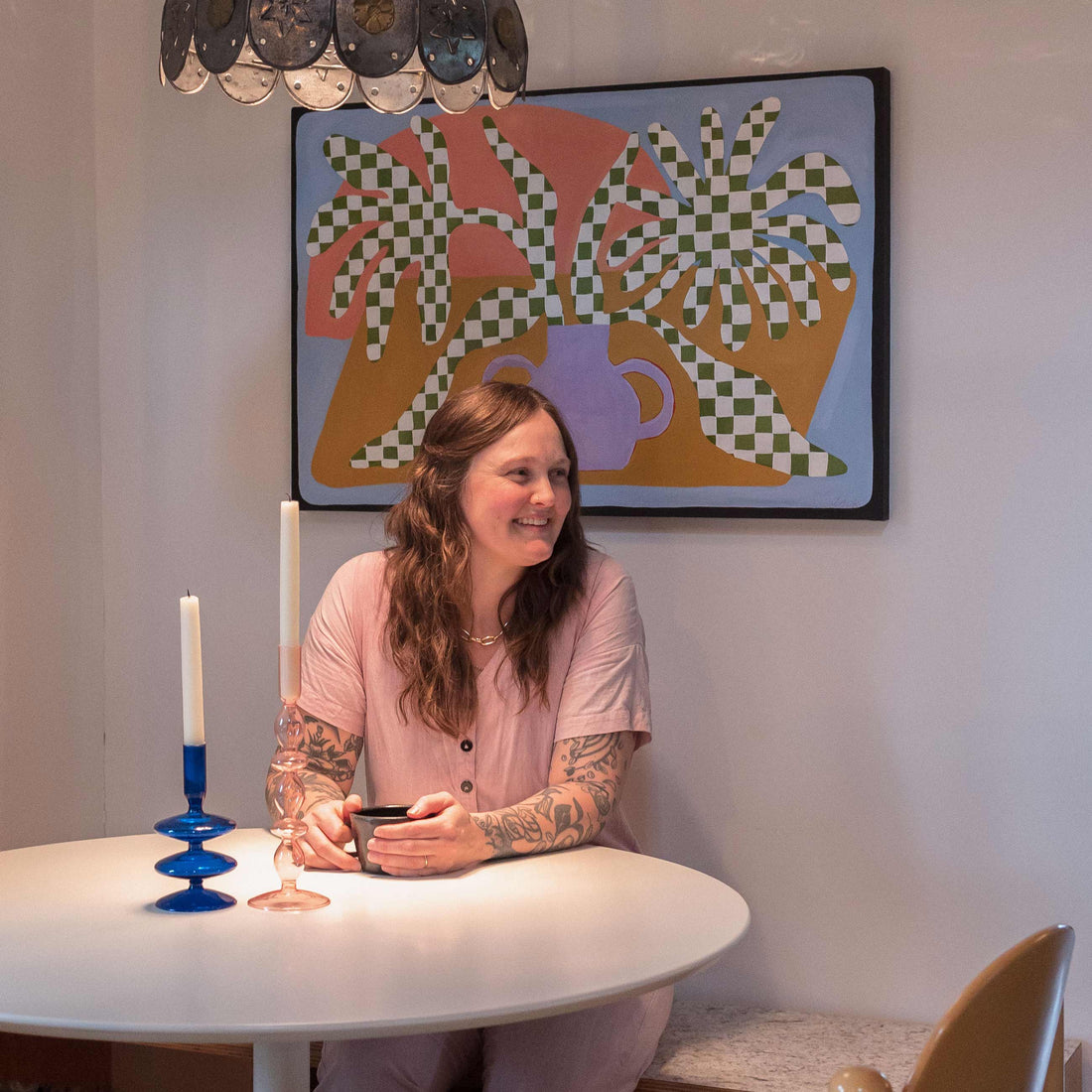 Woman smiling at a round dining table holding a mug, tattoos visible, colorful framed painting and two candlesticks on the table