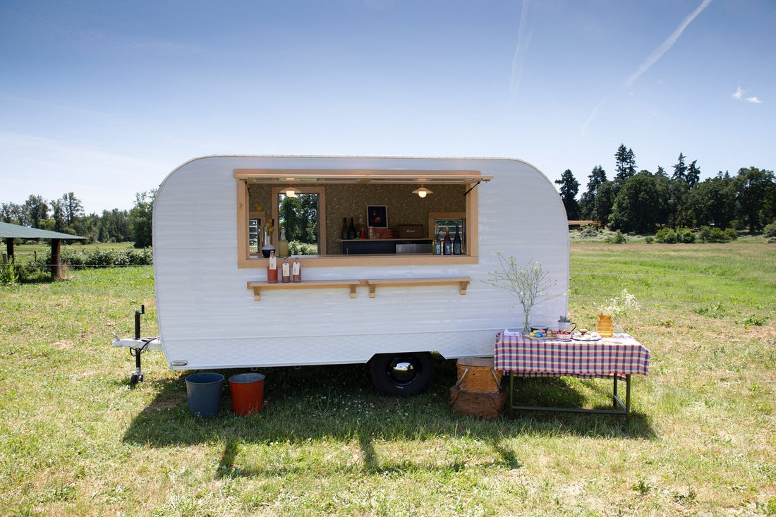White vintage trailer serving drinks in a sunny grassy field, window counter with bottles and a checkered snack table to the right
