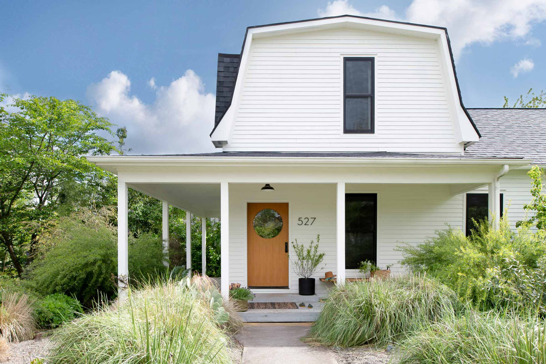 Front porch of white two-story house with covered entry, wooden door with round window, black-trim windows, and ornamental grasses