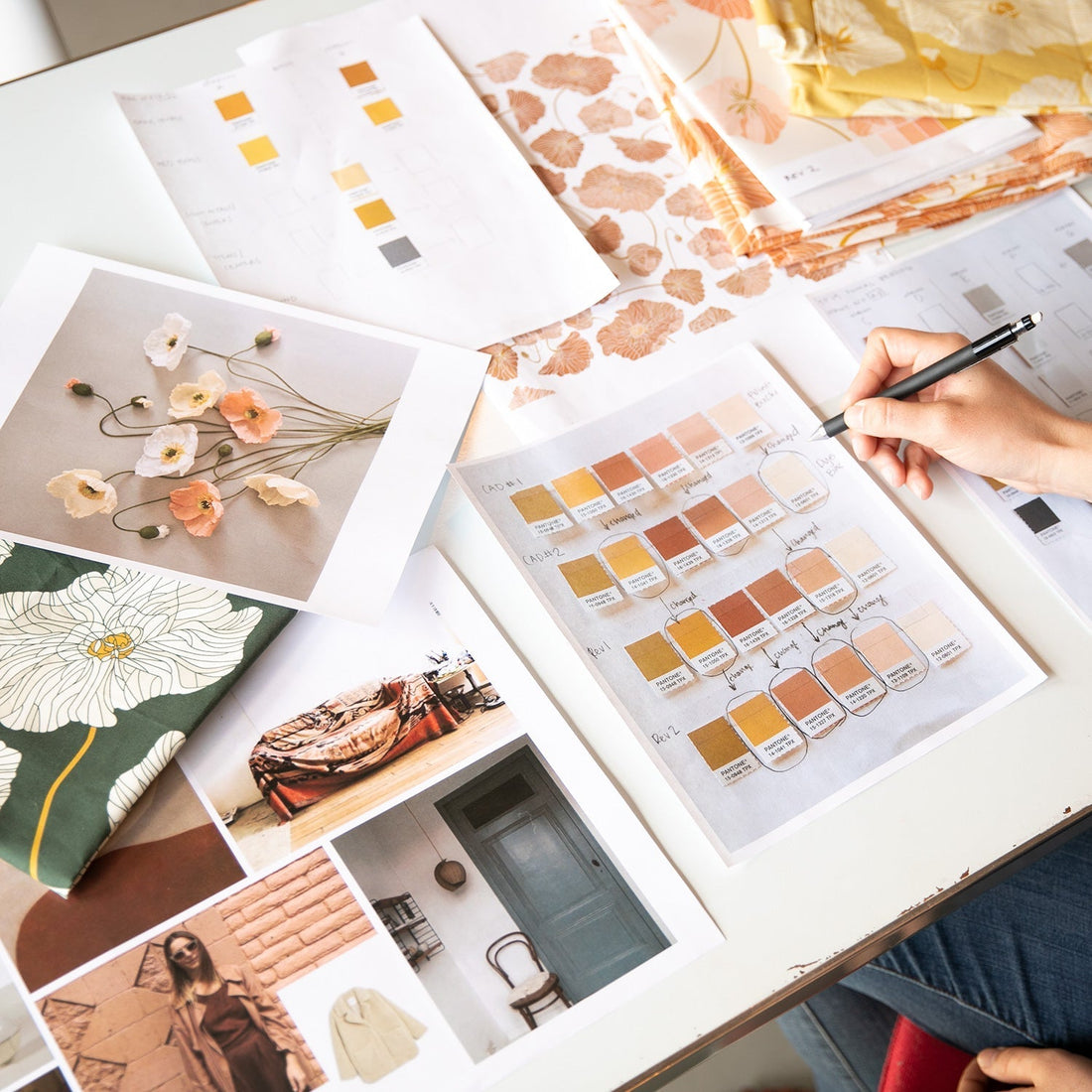 Designer marking warm-toned Pantone color swatches on a white table amid floral pattern prints and fabric samples