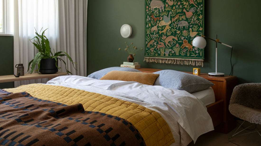 Bed with mustard quilt and patterned brown throw in cozy bedroom with green walls, textile wall hanging and bedside lamp