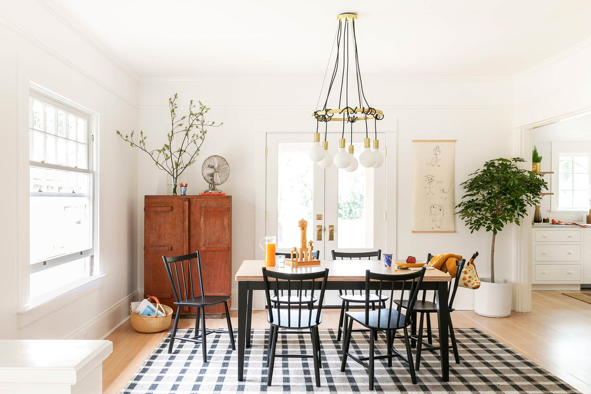 Bright, tidy dining room with wooden table and six black spindle chairs on a black-and-white plaid rug, brass multi-bulb pendant, red storage cabinet, and potted tree