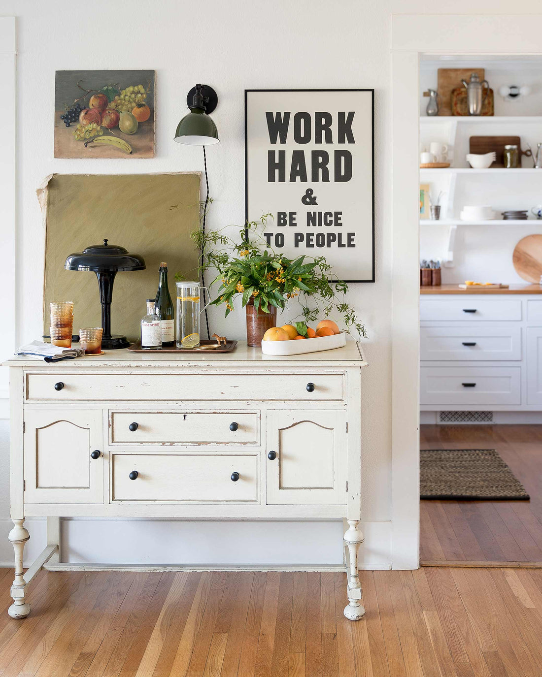 Console table with a green plug-in wall sconce and framed print reading ‘WORK HARD & BE NICE TO PEOPLE’, plant, pitcher, and bowl of citrus