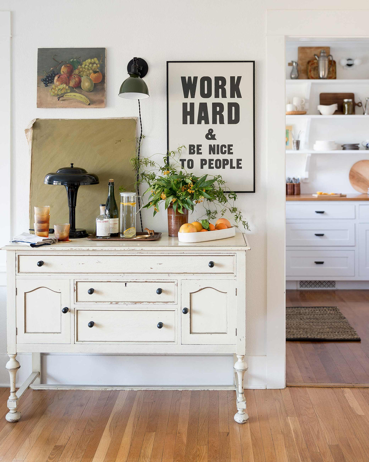 Console table with a green plug-in wall sconce and framed print reading ‘WORK HARD & BE NICE TO PEOPLE’, plant, pitcher, and bowl of citrus