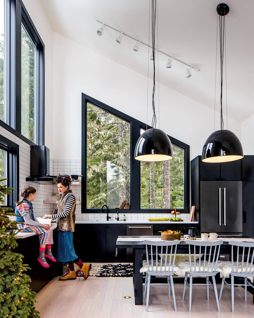 Woman and child chatting in a modern black-and-white ski-chalet kitchen with large triangular windows overlooking a forest