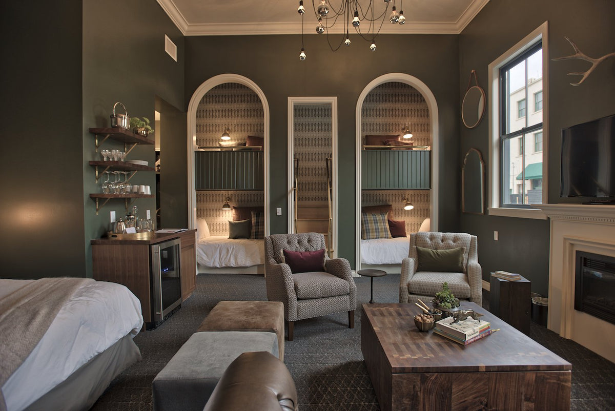 Hotel suite sitting area with two built-in arched bunk beds, upholstered armchairs, wooden coffee table, and deep green walls with a street-facing window