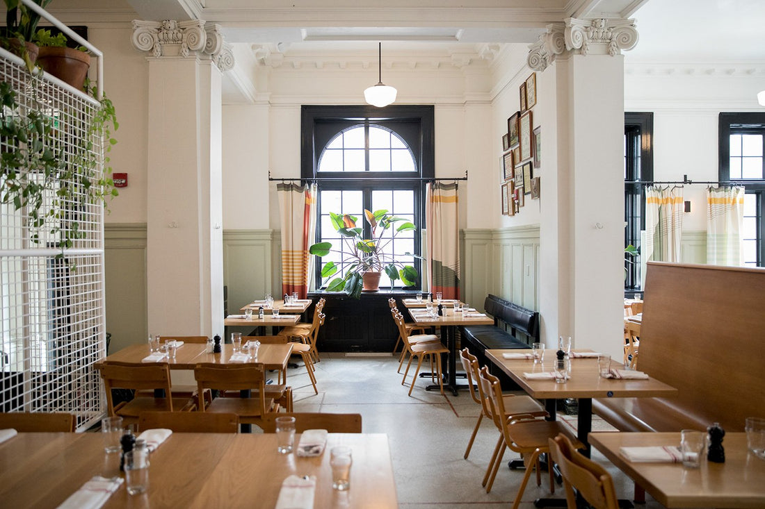 Dining room with wooden tables set for service, arched window and large potted plant at Whitfield in Ace Hotel Pittsburgh