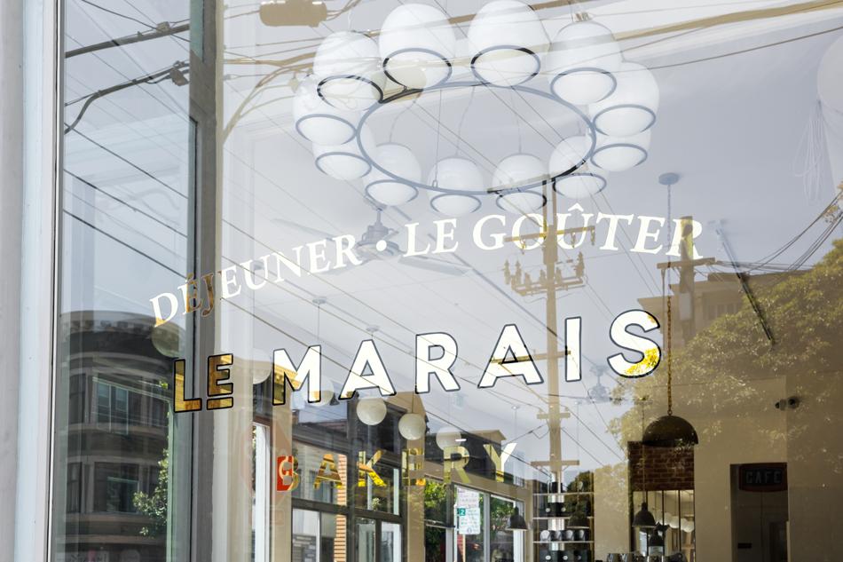 Le Marais Bakery storefront window with gold-lettered signage reading LE MARAIS BAKERY, reflecting the street and showing interior pendant lights