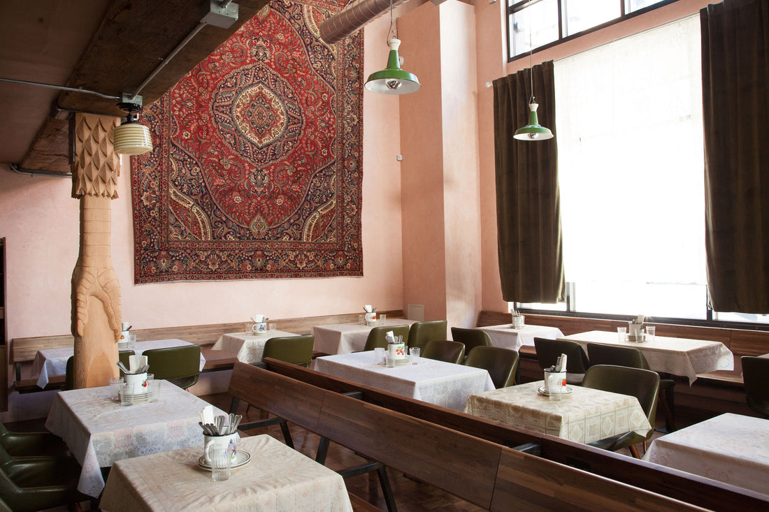 Sunlit restaurant dining room with white-clothed tables, green chairs, a large red Persian rug on pink walls, and pendant lights