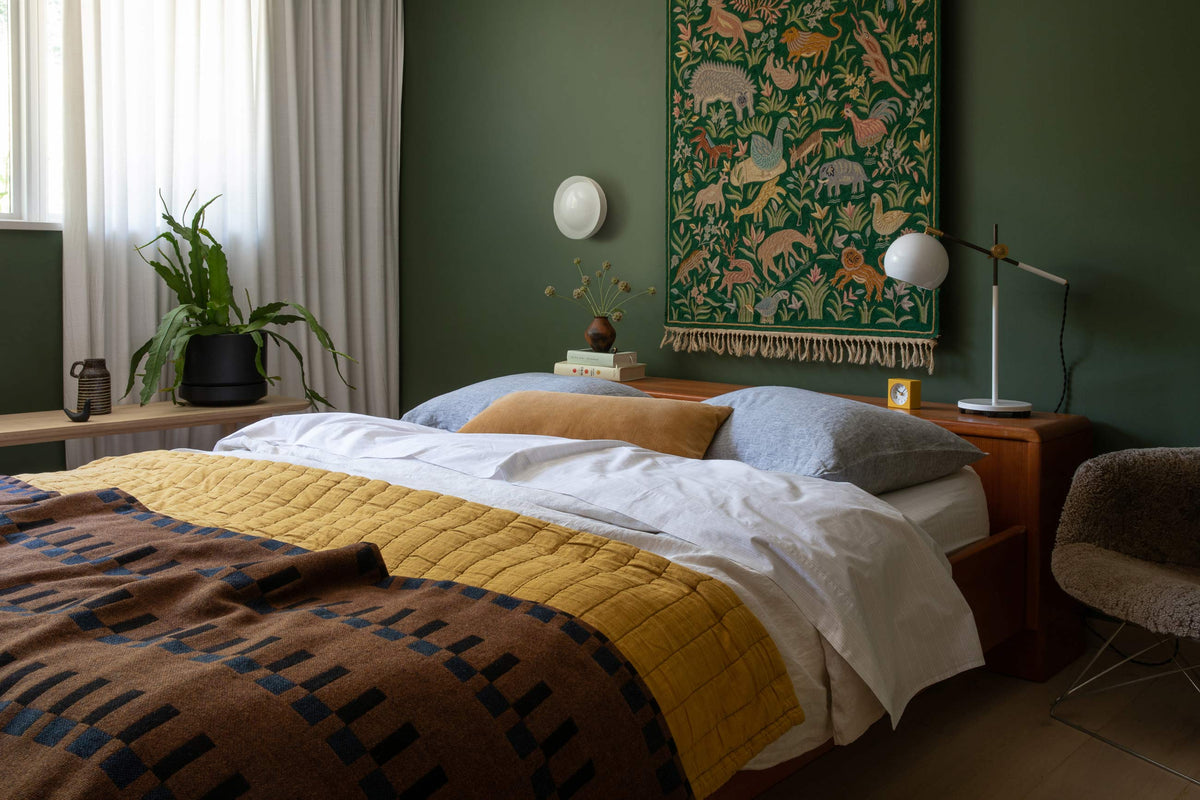 Bed with mustard quilt and patterned throw beneath a white round wall sconce on a deep green wall, wooden nightstand with lamp and tapestry
