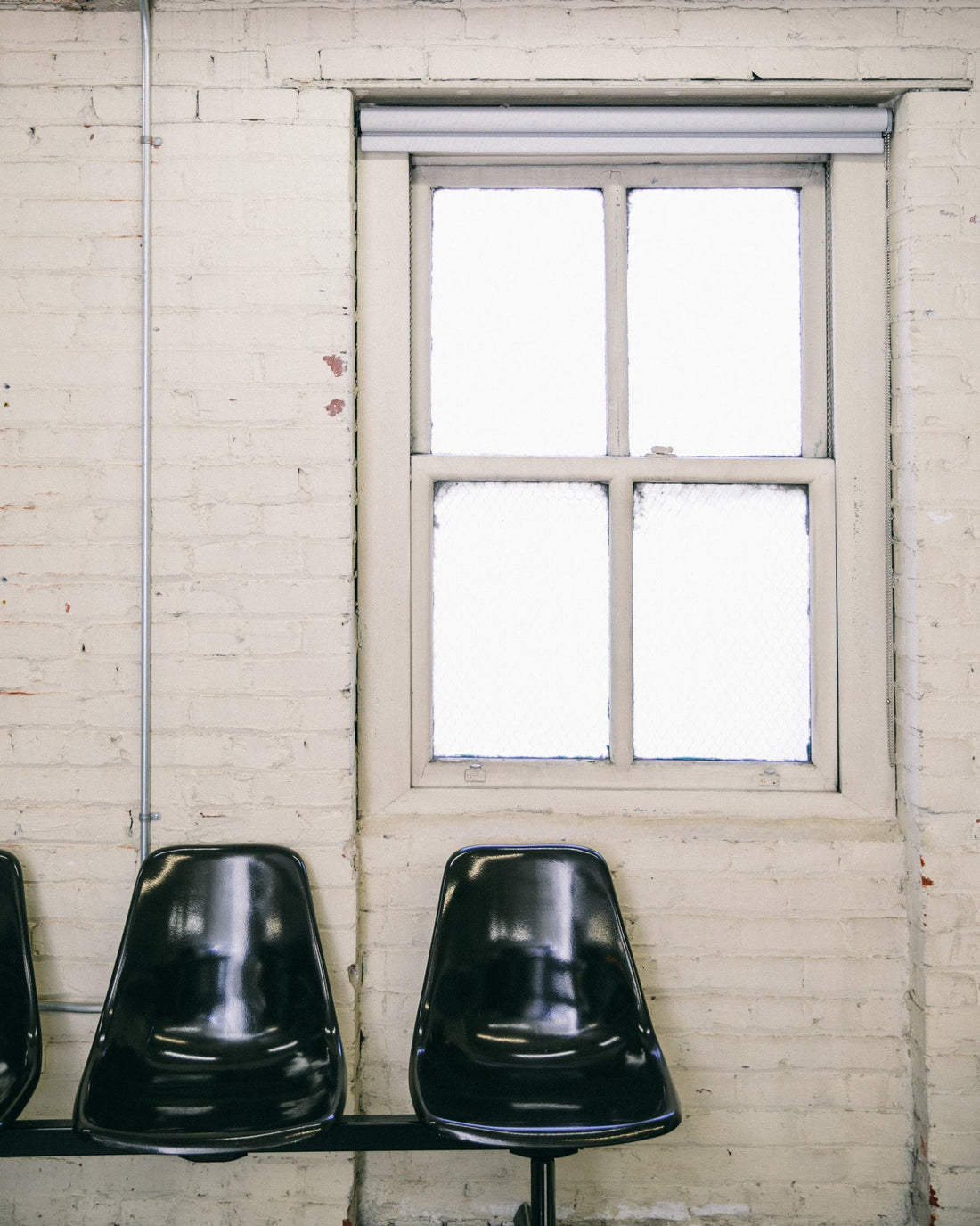 Row of black molded waiting-room chairs under a frosted four-pane window on a painted white brick wall