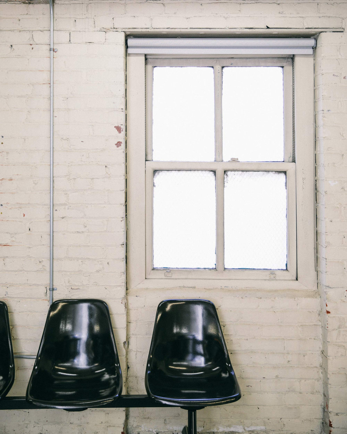 Row of black molded waiting-room chairs under a frosted four-pane window on a painted white brick wall