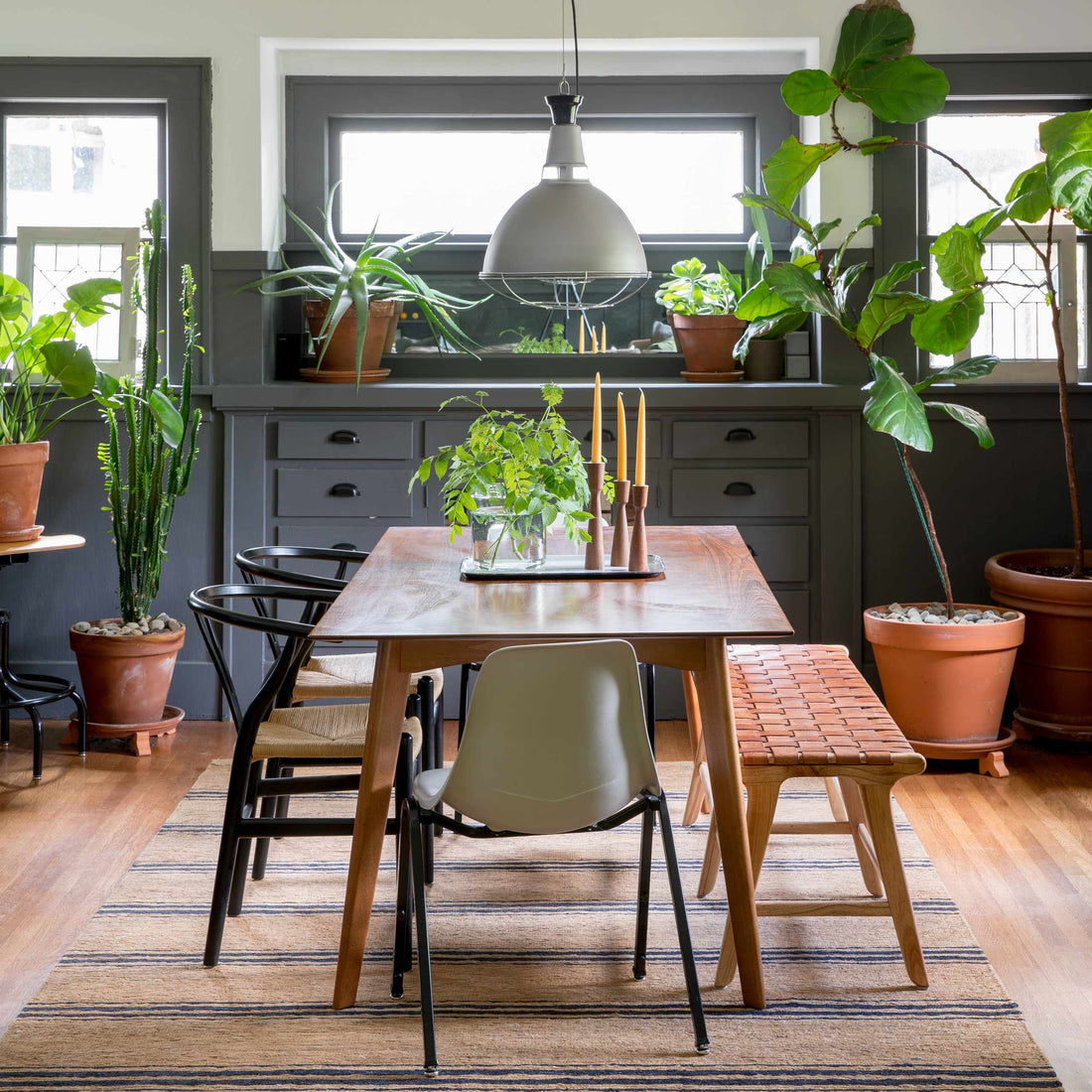Wood dining table with candles and a glass vase plant under an industrial pendant, mixed chairs and potted plants by gray built-in cabinets