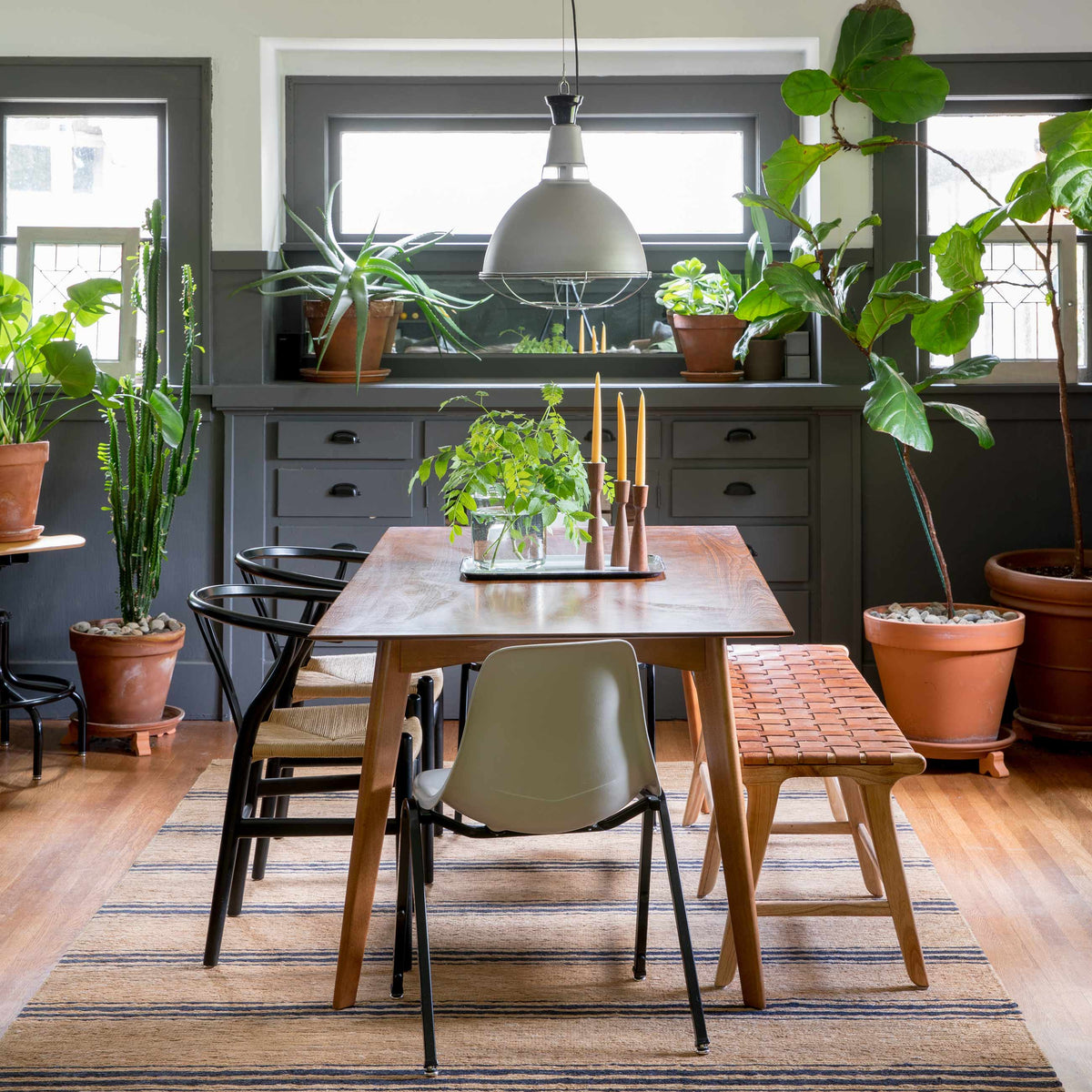 Wood dining table with candles and a glass vase plant under an industrial pendant, mixed chairs and potted plants by gray built-in cabinets