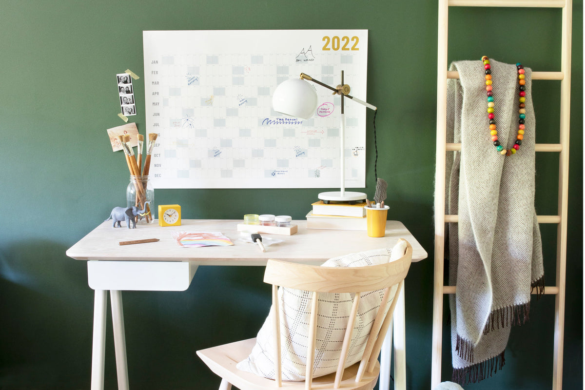 Home desk under 2022 calendar with lamp, paint brushes, alarm clock, and wooden ladder holding a blanket against a dark green wall