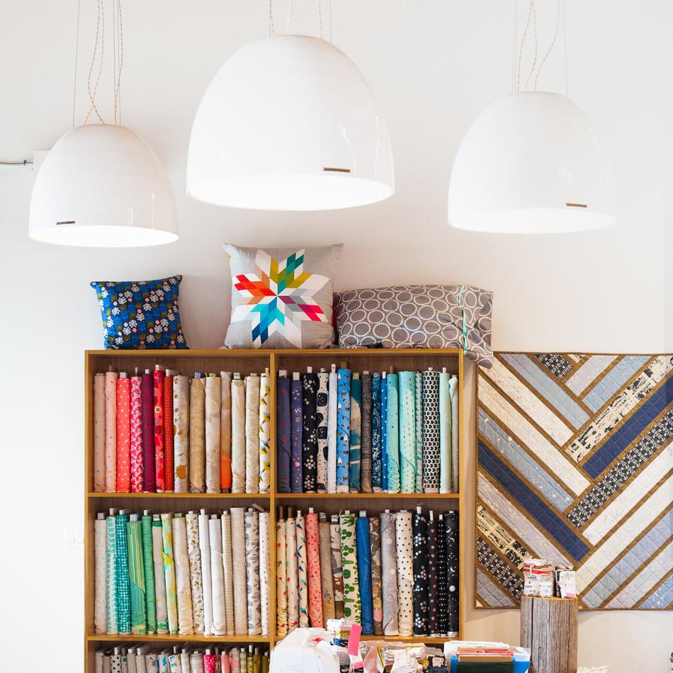 Fabric shop interior with shelves of colorful fabric bolts under three large white pendant lights, decorative pillows on top and a geometric quilt on the wall