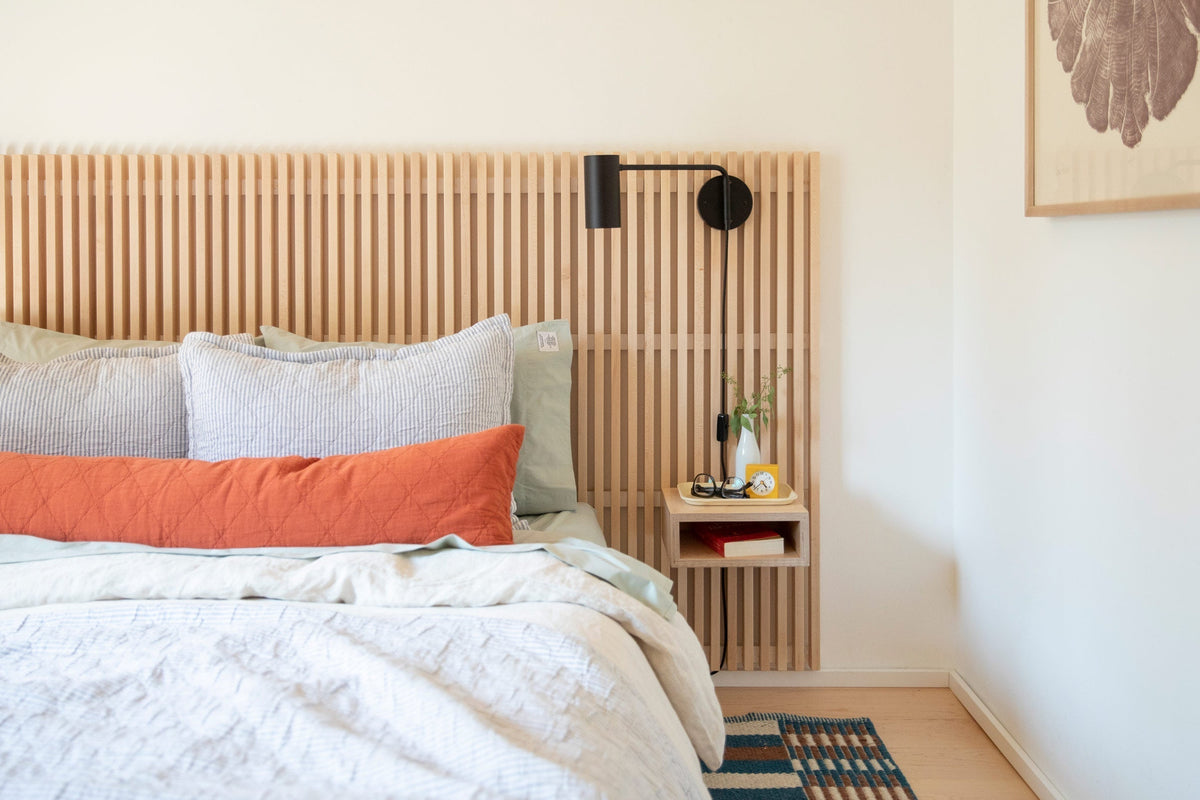 Bed with slatted wood headboard and floating side table styled with black wall lamp, orange lumbar pillow, and small vase