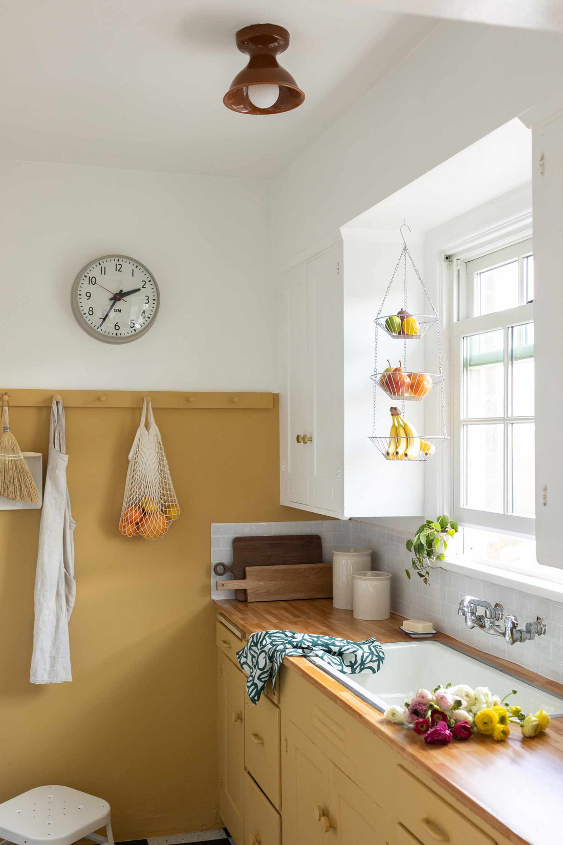 Sunlit kitchen corner with pale yellow cabinets, butcher block counter, hanging three tier fruit baskets, and a wall clock