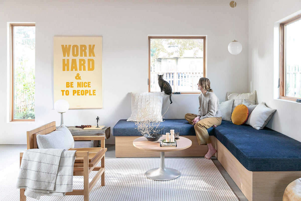 Woman seated on built-in navy bench in a bright living room watching a cat on the windowsill; wall poster reads WORK HARD & BE NICE TO PEOPLE