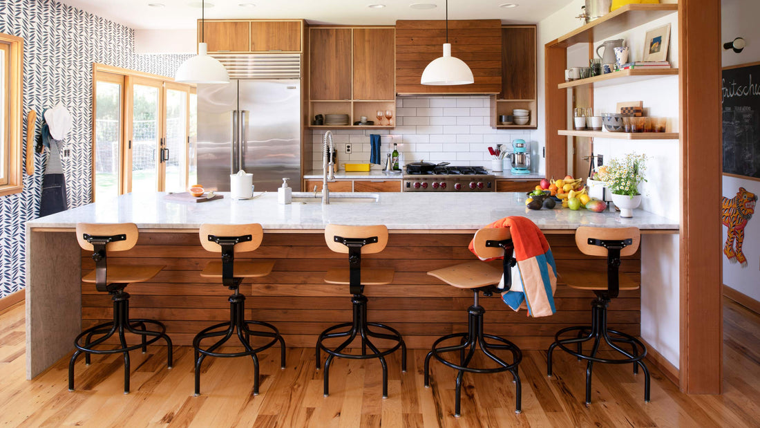 Sunlit kitchen with marble island and five wooden swivel stools, a colorful throw draped over one stool and a bowl of fruit on the counter