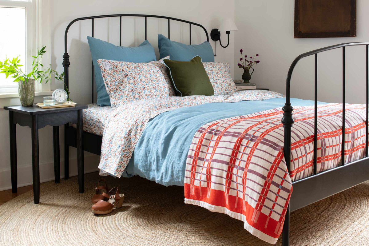 Three-quarter view of coverlet showing red-orange plaid border and quilting on a bed with blue sheets and black iron frame