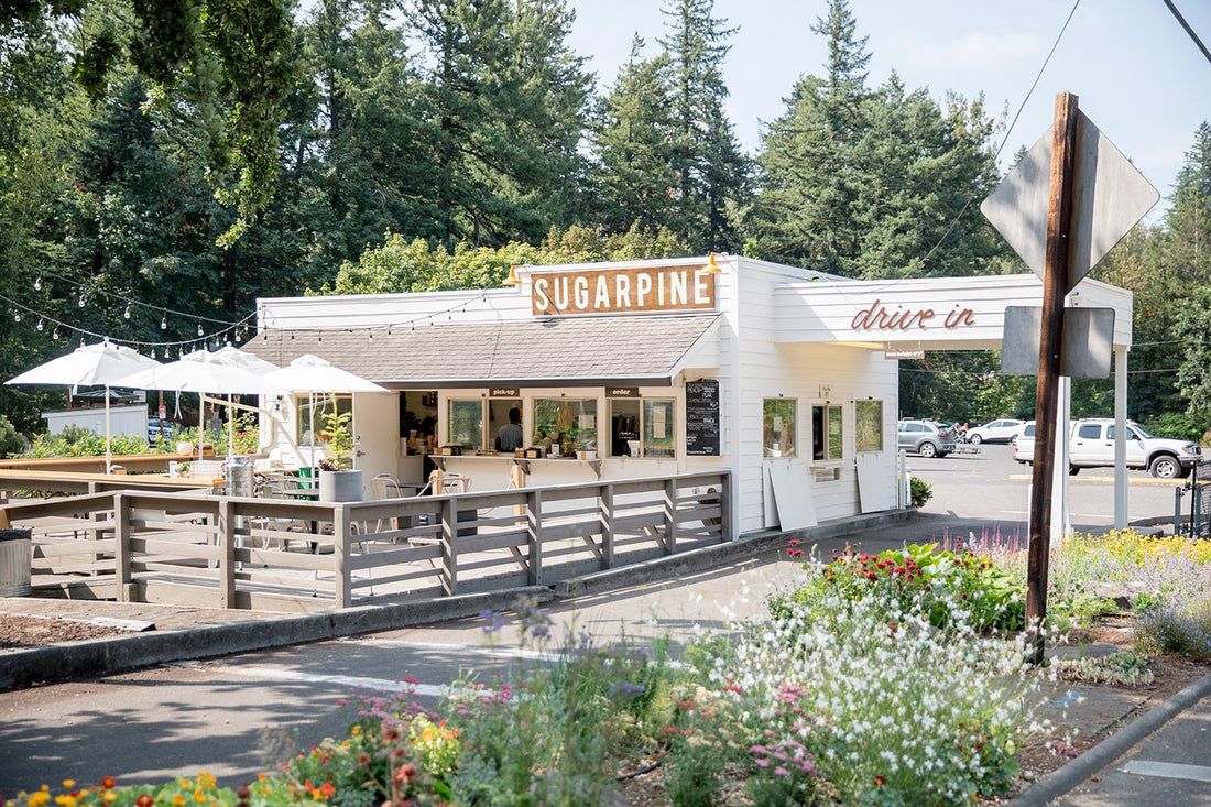 Sugarpine Drive-In building with open service windows, outdoor patio umbrellas, string lights, and flower beds
