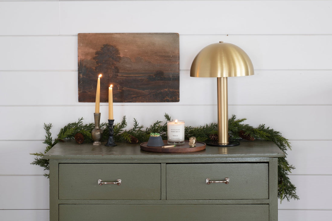 Dresser top with a lit Wildwood jar candle, two taper candles in vintage holders, and a brass dome lamp against white shiplap