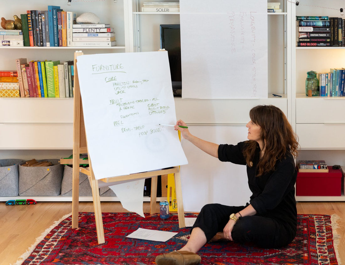 Woman sitting on a rug pointing to a flip chart labeled Furniture during a product development offsite in a bookshelf-lined office
