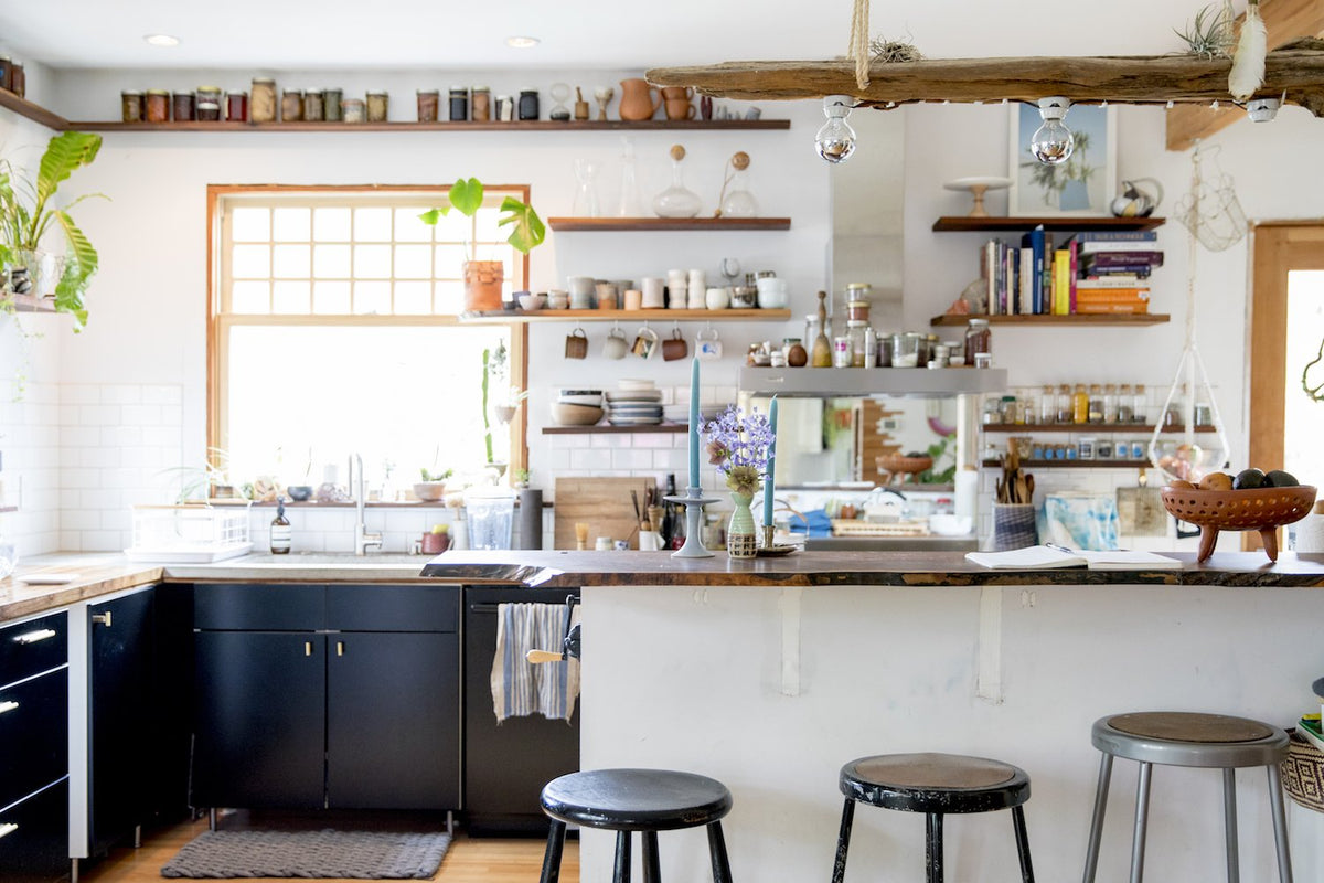 Bright airy kitchen featuring black lower cabinets, open wood shelves, a wooden island with three stools and a vase of purple flowers