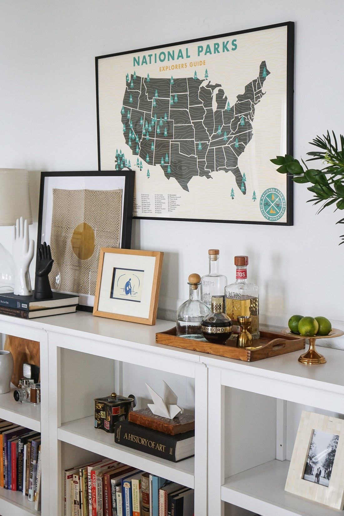 Framed National Parks map hangs above a white console styled with framed art, glass bottles on a wooden tray, and a small potted plant