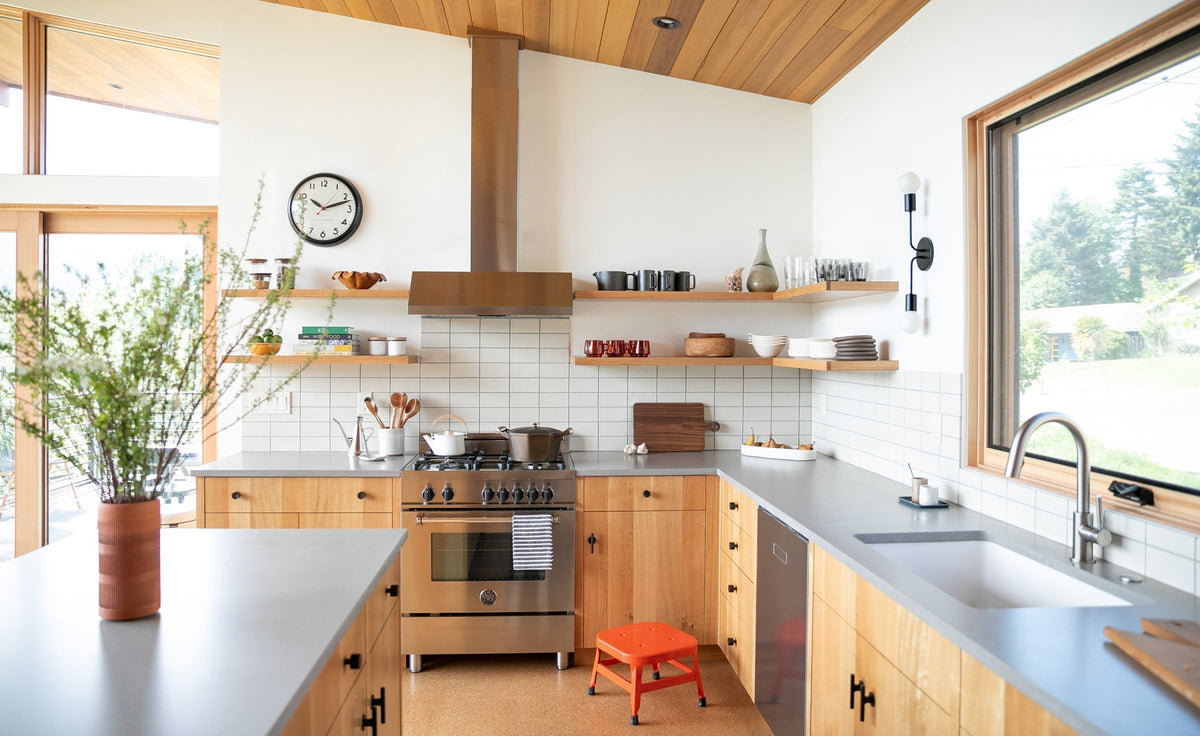 Open kitchen with wood cabinets showcasing a stainless steel range, floating shelves with dishes, and a large window showing greenery