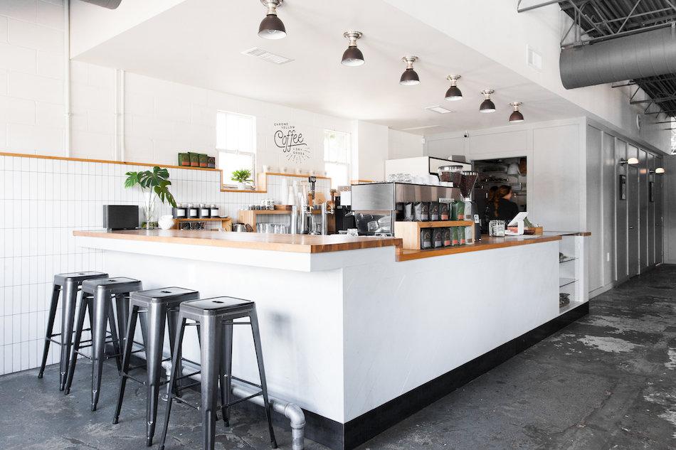Bright minimalist coffee shop counter with wood countertop, espresso machine, four metal stools, white tiled walls, and pendant lights