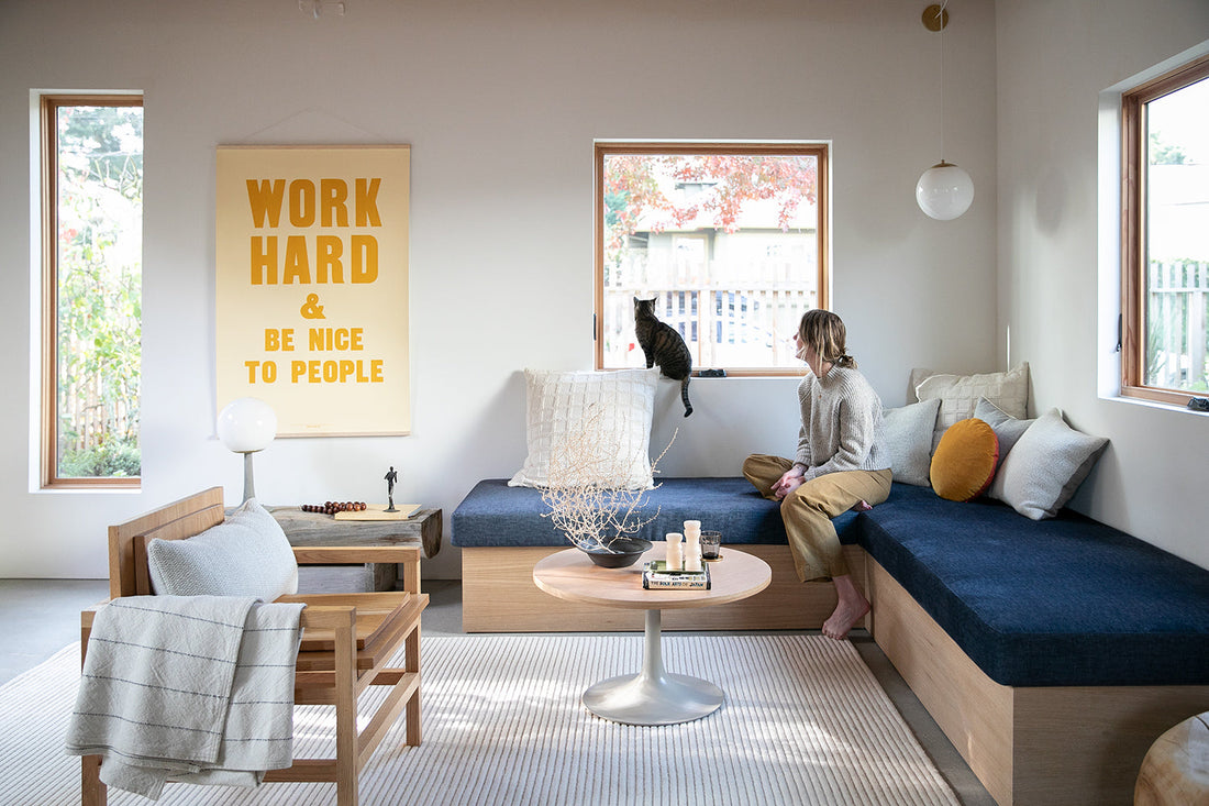 Woman on built-in bench watching a cat on a windowsill in a bright living room with cushions and a yellow poster reading WORK HARD AND BE NICE TO PEOPLE