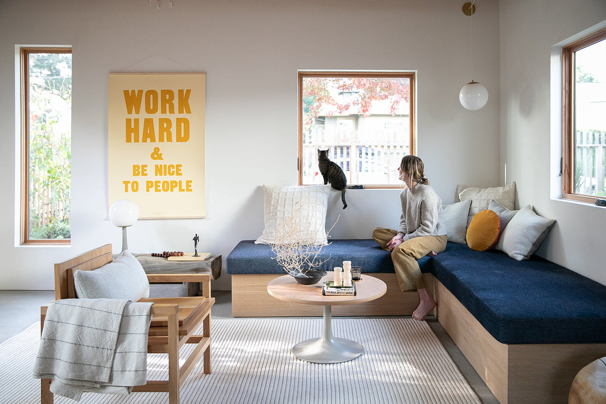 Woman on built-in bench watching a cat on a windowsill in a bright living room with cushions and a yellow poster reading WORK HARD AND BE NICE TO PEOPLE