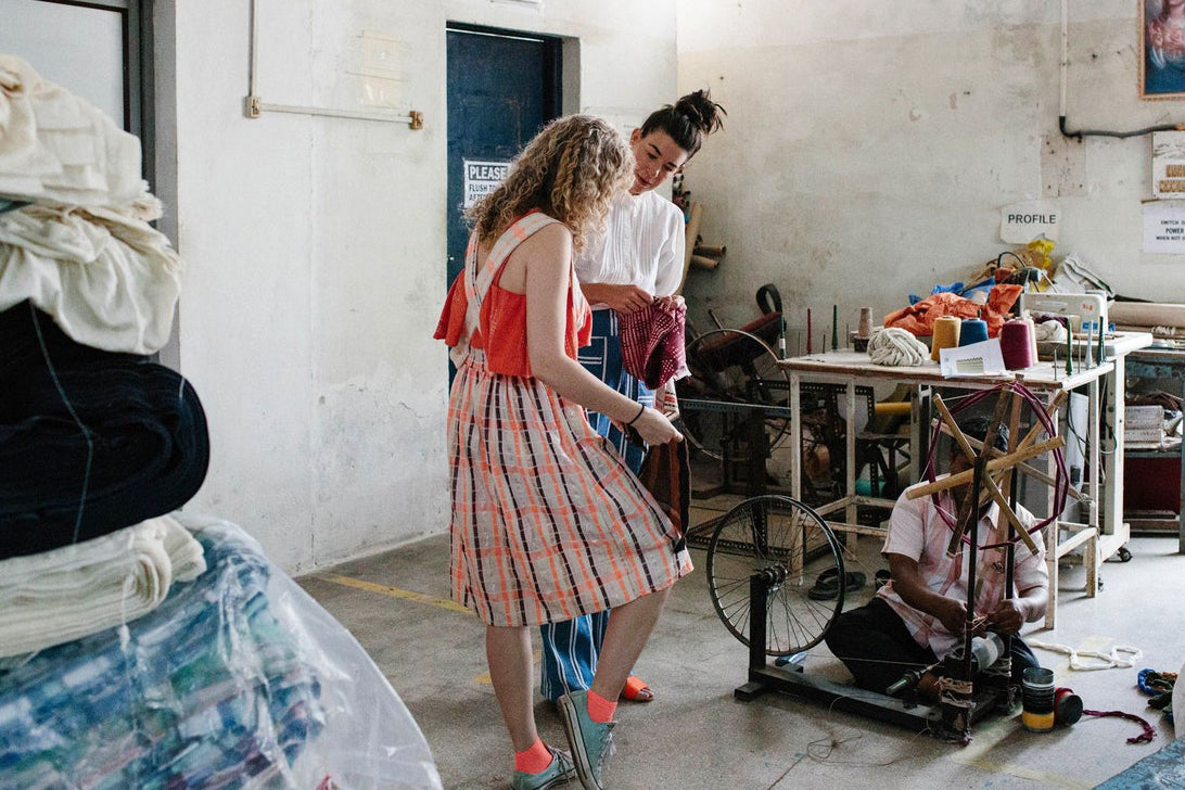 Two designers examining woven fabrics in a textile workshop with a spinning wheel and stacks of cloth