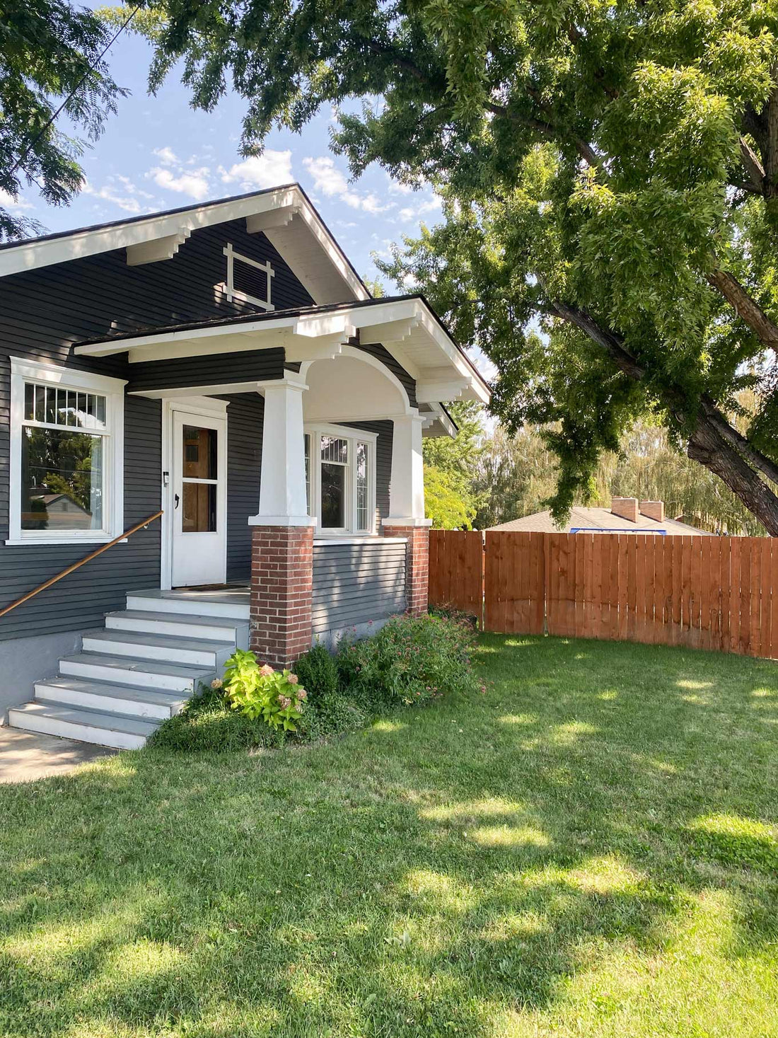 Front exterior of a navy-gray Craftsman house with white trim and brick porch columns, shaded by a large leafy tree
