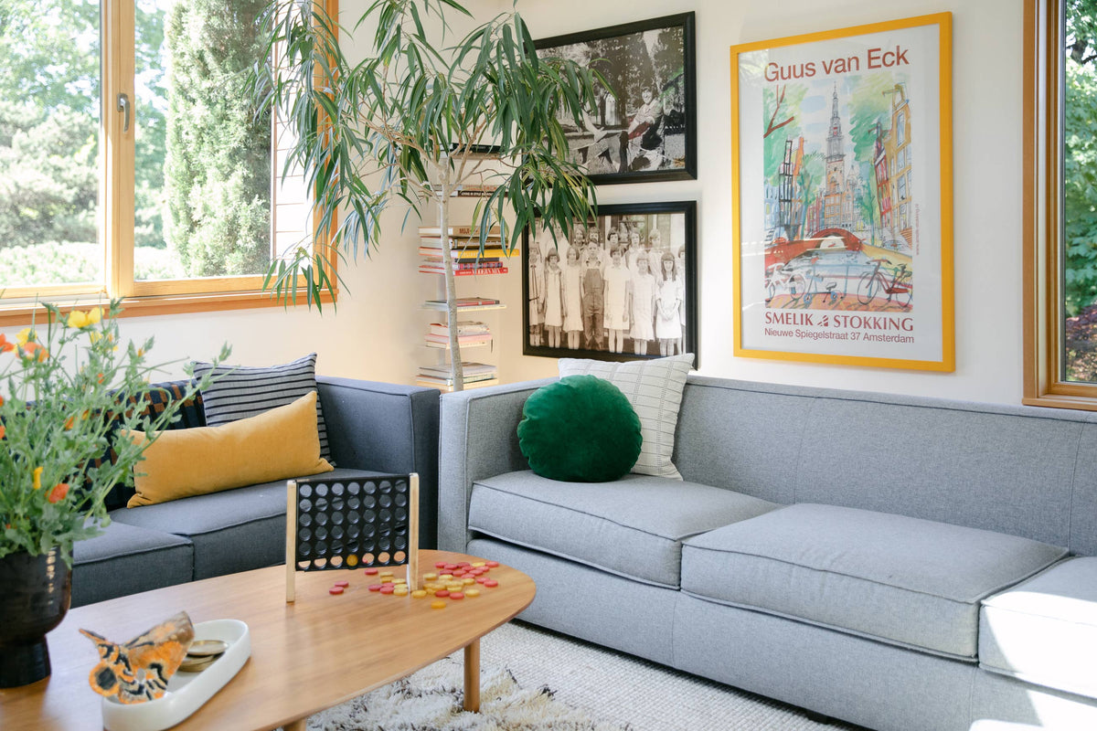 Sunlit living room with gray sectional sofa, framed photos and colorful poster, coffee table with game pieces and plants