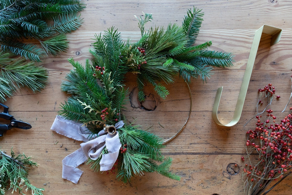 Half-finished evergreen winter wreath on a gold hoop being assembled on a wooden table with red berries, ribbon, and shears