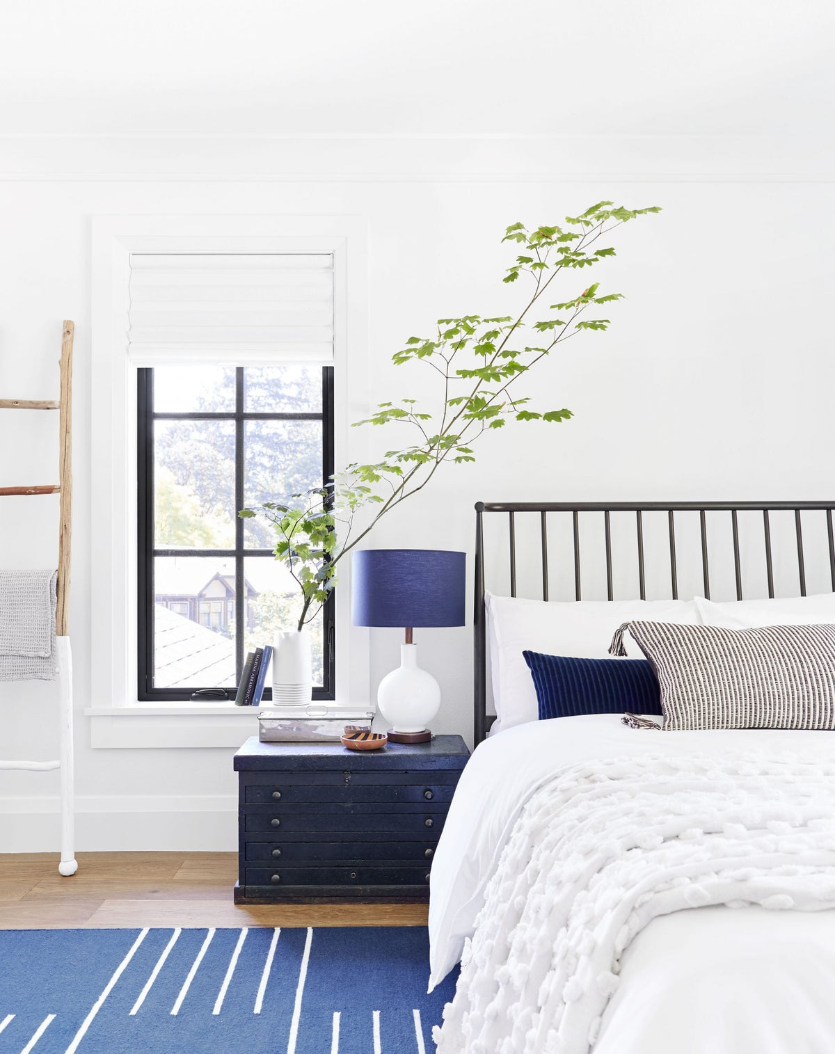 Bedside showing Bond lamp on a navy nightstand with a leafy branch, black metal headboard, white bedding, and blue rug