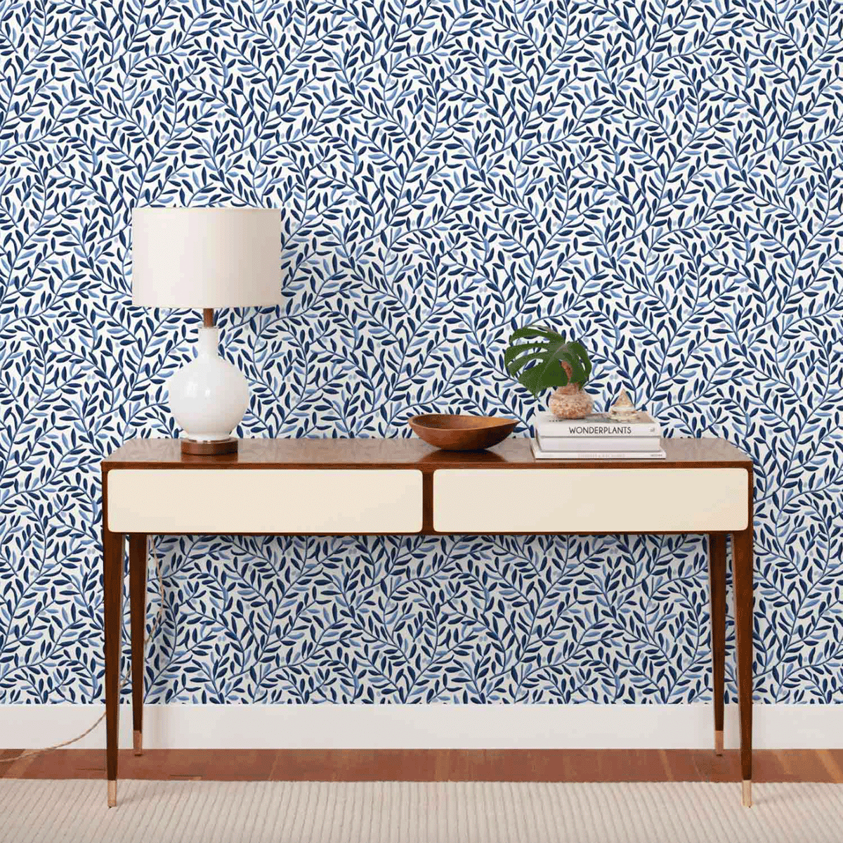 Console table styled with a white ceramic lamp, wooden bowl, and potted plant against blue leafy wallpaper