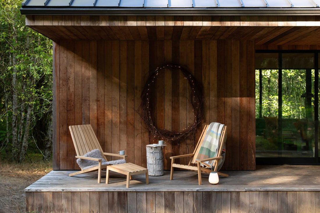 Two wooden lounge chairs flank a log side table on a rustic cabin porch with a lit twig wreath on the wood wall and trees beyond